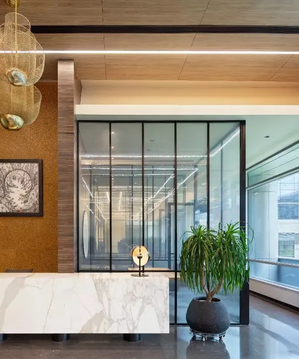 The reception lobby at the APT office in New Delhi, featuring a marble desk and warm wood tones. The design establishes a professional yet inviting first impression, balancing corporate sophistication with natural materials and abundant light from the large windows.