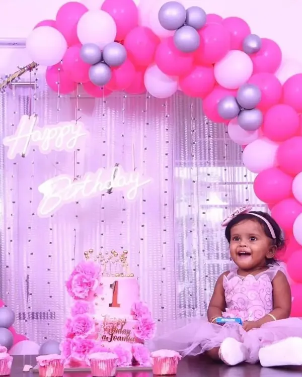 A precious moment from a first birthday party, featuring a beautiful pink and white balloon arch against a shimmer curtain backdrop. The setup includes a custom cake and a "Happy Birthday" neon sign.