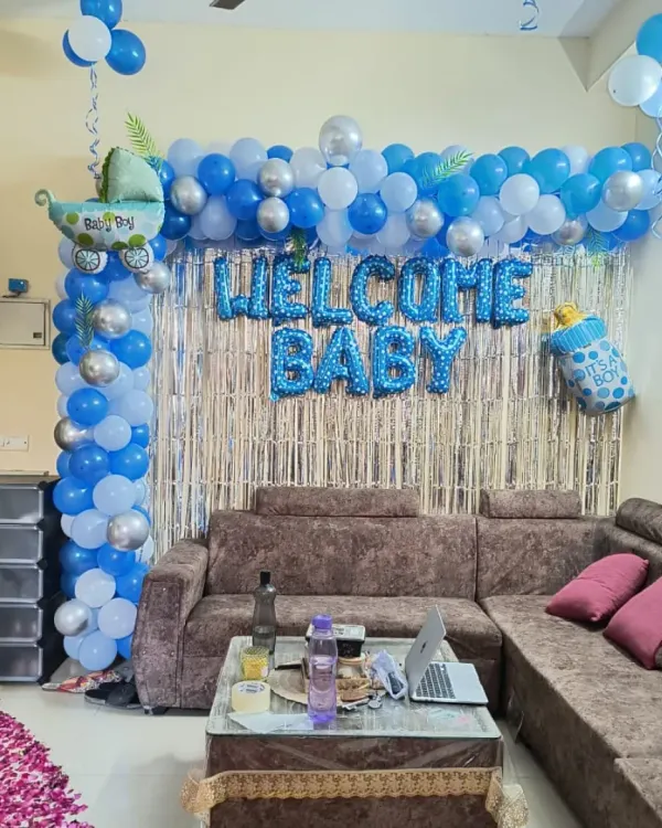 A 'Welcome Baby' decoration for a baby boy in a living room. The setup includes a blue and white balloon frame, a silver shimmer curtain, and baby-themed foil balloons.