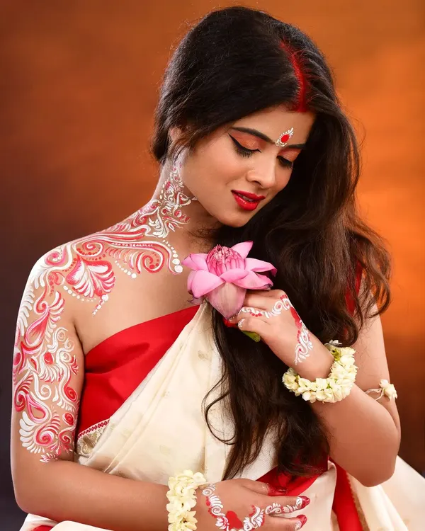 A traditional Bengali look for Durga Puja, featuring intricate, hand-painted Alpona on the neck, shoulder, and arm. This body art complements the classic red and white saree, creating a complete, culturally rich character.