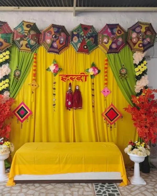 A traditional Haldi stage featuring a bright yellow backdrop, a line of decorative Rajasthani umbrellas, and hanging puppets. The setup is framed with marigold garlands and accented with colorful artificial foliage for a full, festive look.