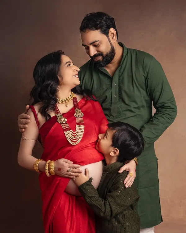 A beautiful family maternity portrait rich with emotion. The expecting mother in a vibrant red saree shares a loving gaze with her husband as their son kisses her baby bump.
