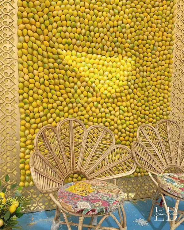 A stunning Haldi seating area featuring a backdrop made entirely of fresh lemons. The natural cane peacock chair with a colorful cushion adds a bohemian touch to this vibrant, Positano-inspired design.