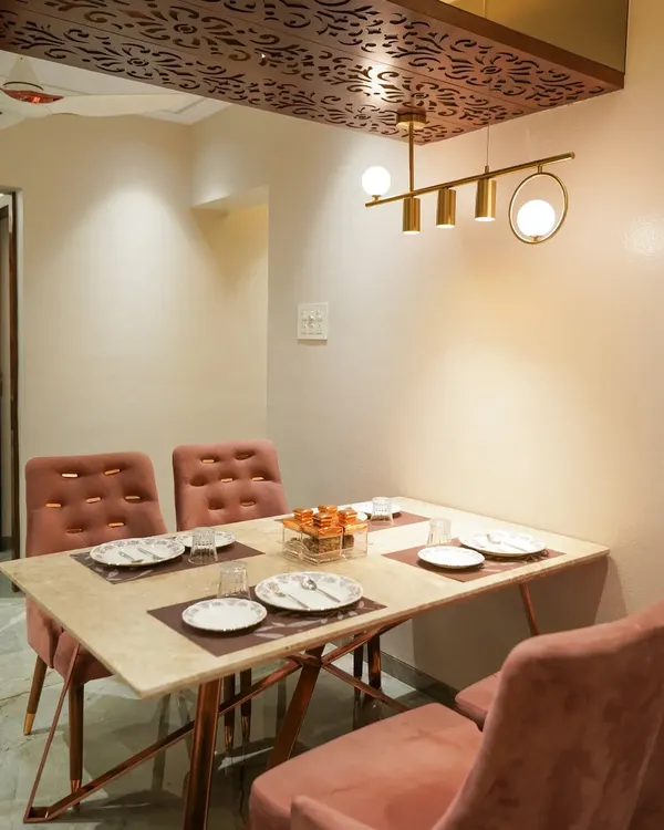 A cozy and elegant dining area. The space features a marble-top table with rose gold legs, comfortable upholstered chairs, and a decorative wooden ceiling panel with a modern chandelier.