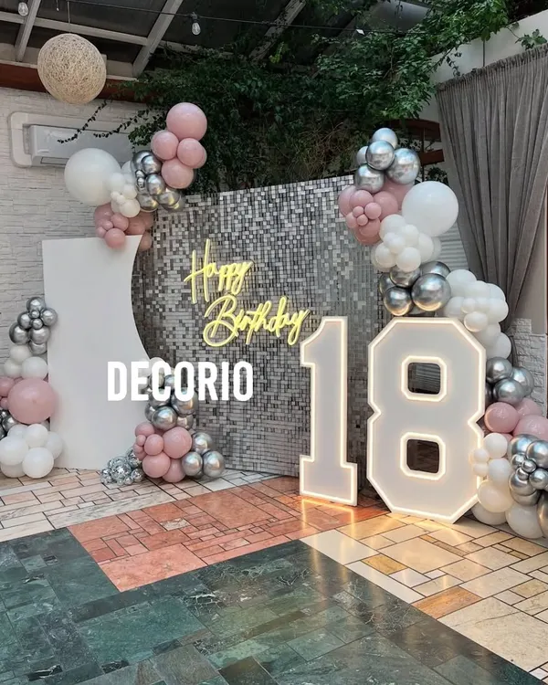 A glamorous 18th birthday setup featuring a silver shimmer wall, a 'Happy Birthday' neon sign, and large marquee numbers. The pink, white, and silver balloon arrangement adds a soft and celebratory touch.