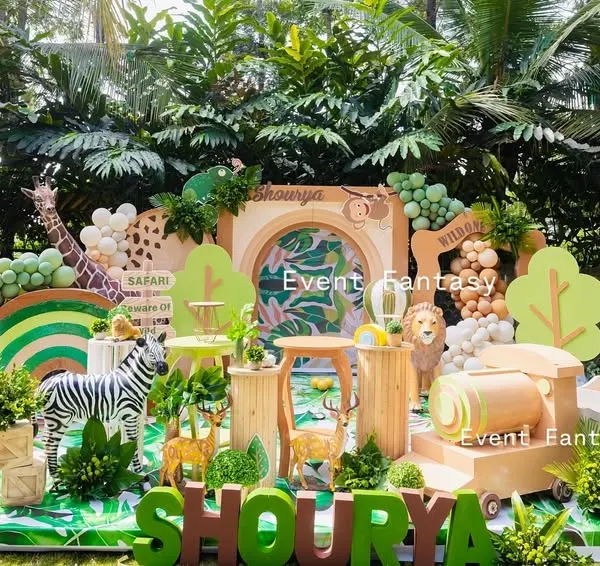 A complete safari adventure setup for Shourya's first birthday. This wide-angle view showcases a lush green outdoor space transformed with a large backdrop, a variety of animal props including a gorilla and zebra, and a custom name display.