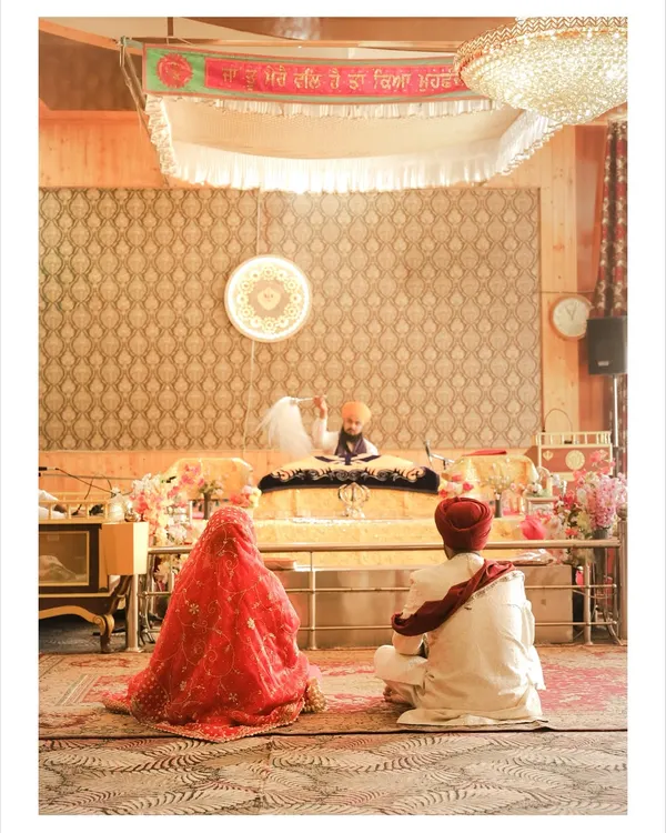A wide shot of the Anand Karaj ceremony inside the Gurudwara. The couple is seen seated before the Guru Granth Sahib, capturing the spiritual essence of a Sikh wedding.