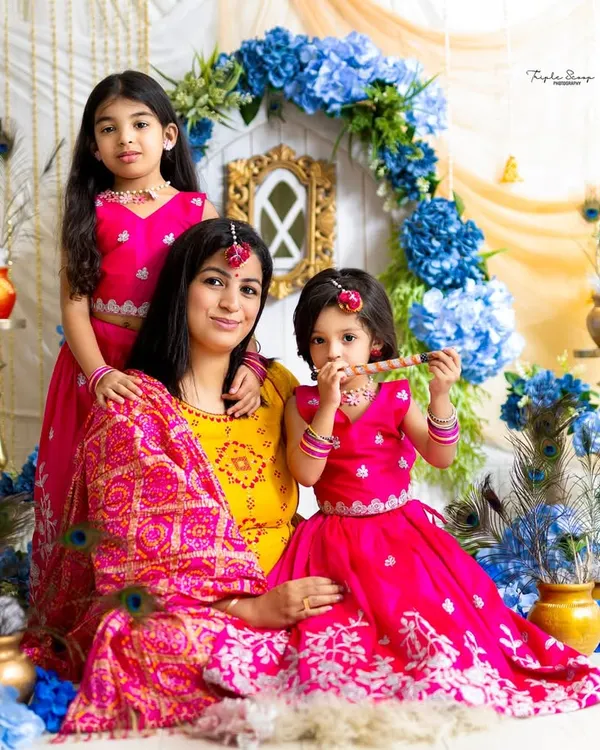 A beautiful Janmashtami-themed portrait of a mother and her two daughters dressed as little Radhas. The vibrant pinks and blues of the set create a festive feel.