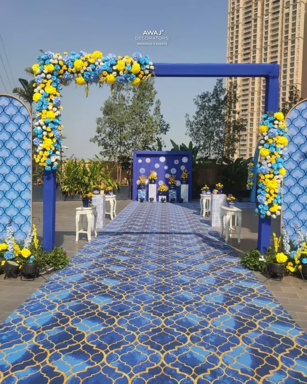 A grand entrance for a Haldi ceremony, featuring a bold blue archway adorned with yellow and blue flowers, leading down a patterned carpet.