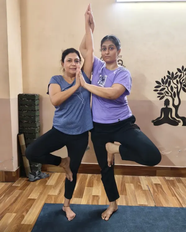 Two students practicing a partner variation of Vrikshasana (Tree Pose). This pose challenges individual balance while relying on a partner for stability, symbolizing mutual support and connection.