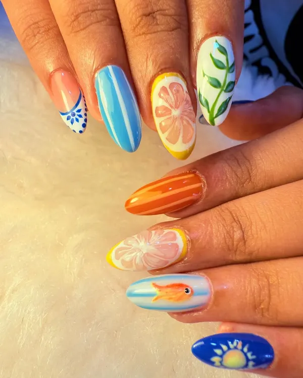 A perfect example of a fun, mismatched summer manicure. Each nail has a unique design, from citrus slices and stripes to a tiny fish and a sunset, all coming together for a playful and creative look.