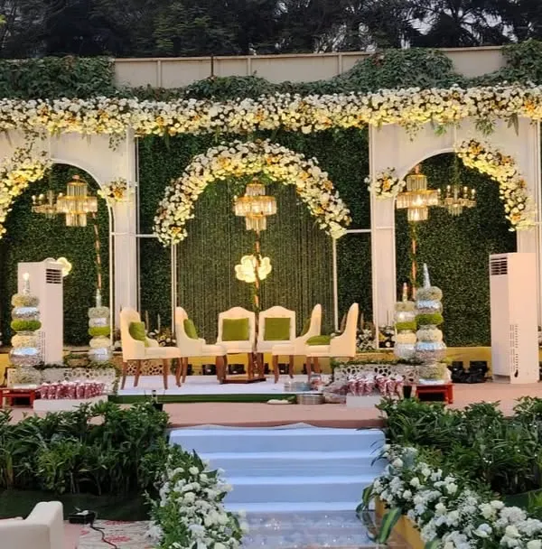 I designed this elegant outdoor reception stage with a backdrop of lush greenery and white floral arches. The setup includes beautiful chandeliers and comfortable seating, creating a sophisticated and romantic ambiance for the evening.