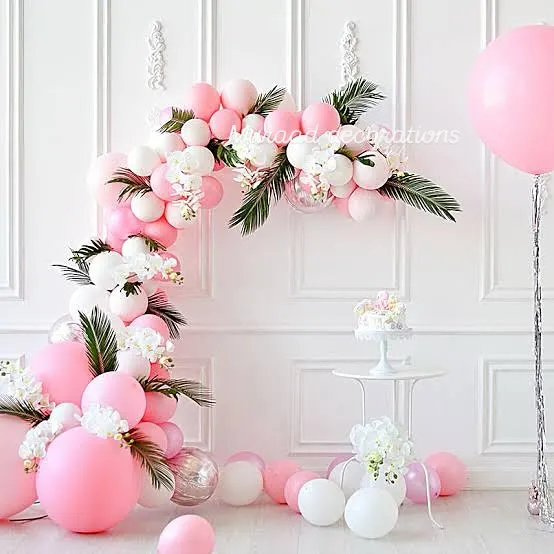 An elegant and airy balloon decoration for a birthday. The pink and white balloon arch is beautifully accented with white flowers and green ferns, creating a fresh and sophisticated look against a clean white wall.