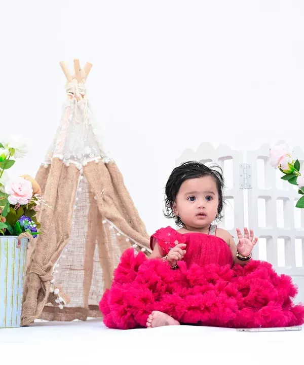 A beautiful baby girl in a vibrant pink dress sits against a clean, white background. This simple setup, with just a few floral and teepee props, keeps the focus entirely on her.