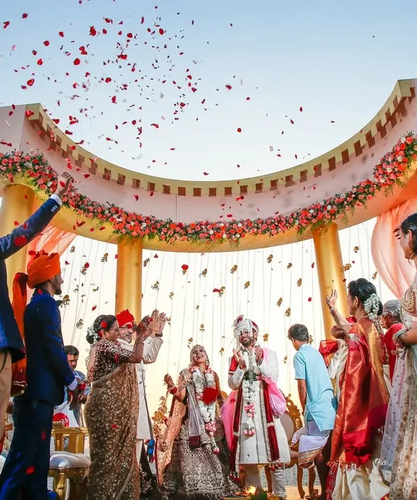 A grand varmala moment under a beautiful arch, with guests showering the couple with rose petals. This wide shot captures the scale and joy of the celebration as the couple officially begins their life together.
