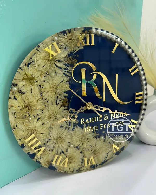 A classic example of our 12-inch custom resin clock, featuring varmala flowers in a moon-shaped design against a shimmery royal blue base. The piece is personalized with a couple's monogram, names, and wedding date in gold.