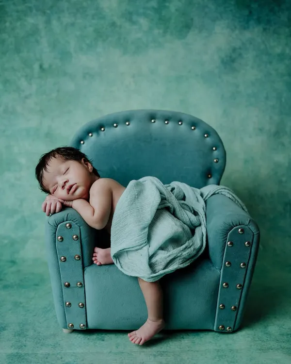 I create calm and artistic settings for newborn photoshoots. Here, a sleeping baby is posed on a miniature teal armchair, with a matching backdrop and soft wrap, highlighting the peaceful and styled nature of my newborn art sessions.