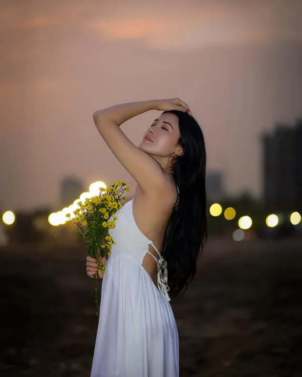 A beautiful beach portrait of a woman in a white dress holding yellow flowers, with the city lights creating a soft bokeh in the background during twilight.