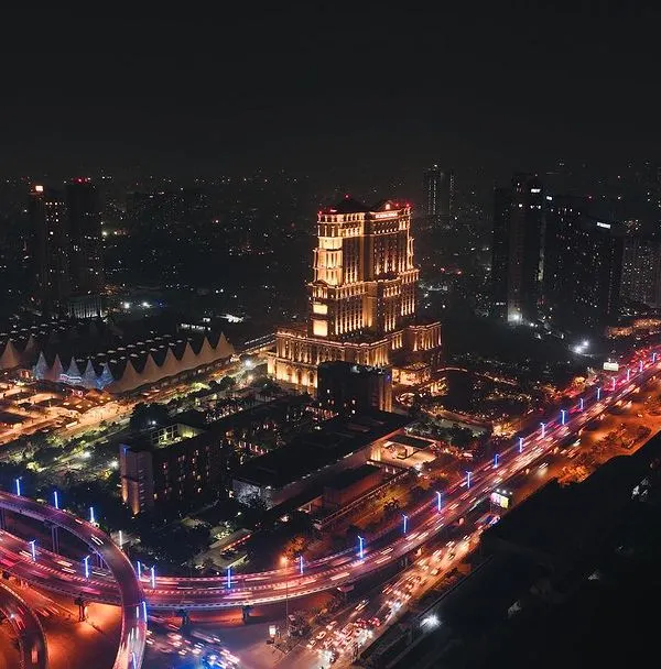 A long exposure aerial photograph of Kolkata at night, capturing the light trails from traffic flowing around the ITC Royal Bengal. This technique transforms a busy city scene into a dynamic and artistic image, showcasing the urban energy from a unique perspective.
