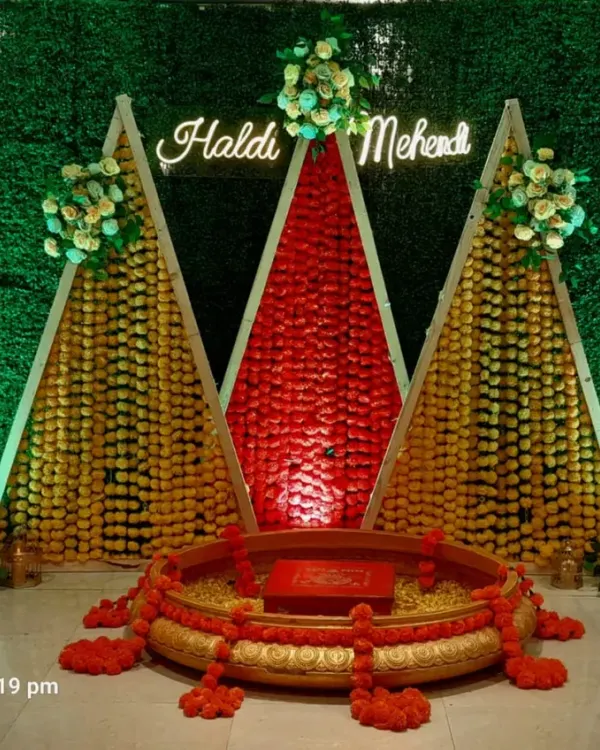 A unique Haldi and Mehendi backdrop featuring a green foliage wall and triangular structures decorated with marigold and jasmine garlands. A neon sign adds a modern touch.