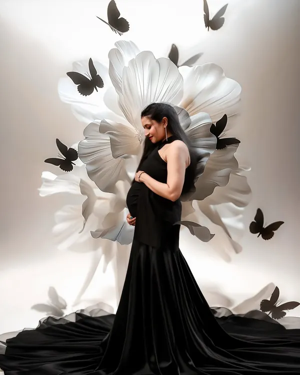 A masterpiece of elegance. The mom-to-be in a stunning black gown is framed by a giant white paper flower and delicate black butterflies, creating a high-fashion, artistic portrait.
