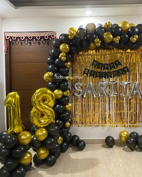 A glamorous 18th birthday decoration in black and gold. This home setup includes a balloon arch, a gold shimmer backdrop, and large foil number balloons.