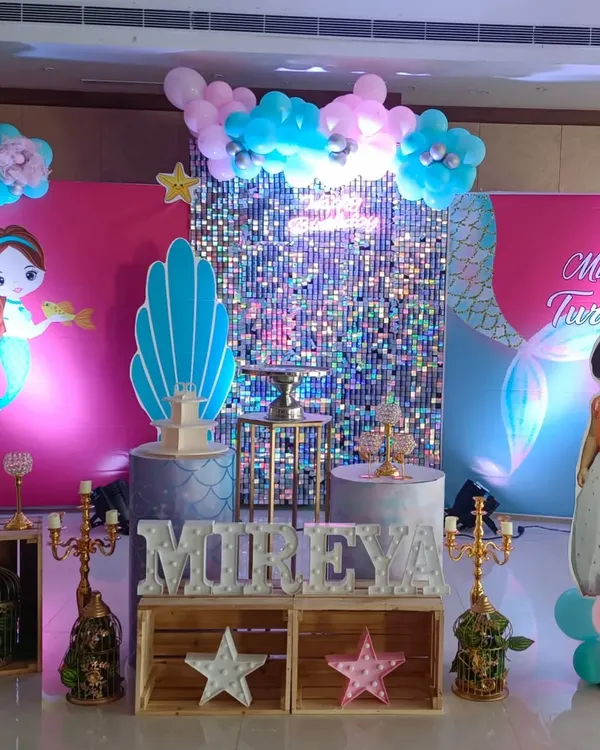 A complete mermaid theme birthday party decoration for a third birthday. This setup includes a silver shimmer wall, custom mermaid and character cutouts, themed pedestals for the cake, and balloon clouds.