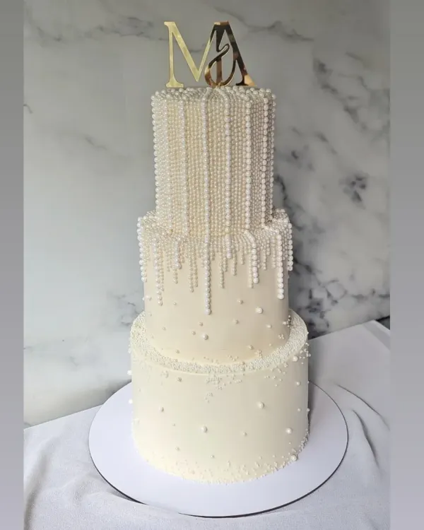 This three-tier wedding cake is a perfect example of timeless elegance. I meticulously placed hundreds of edible pearls to create a cascading waterfall effect, topped with a custom gold monogram for a truly personal touch. It's a non-fondant design with a flawless buttercream finish.