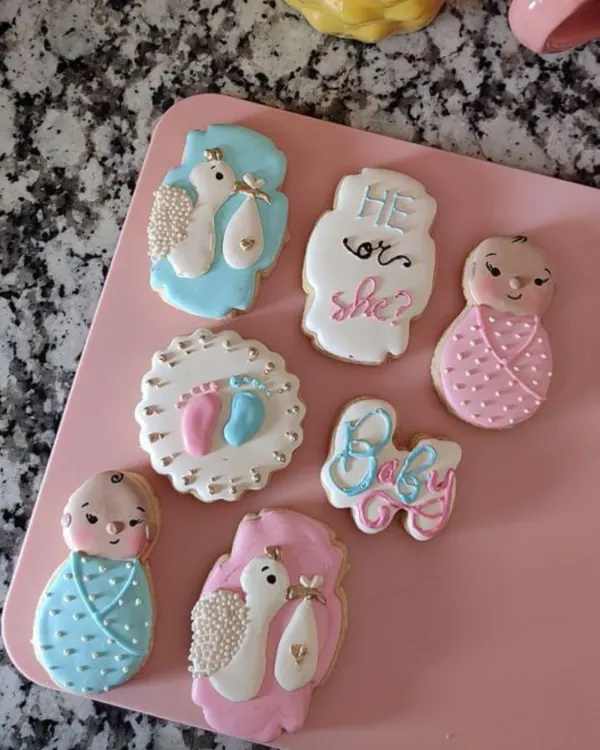 A beautiful set of royal icing cookies made for a baby shower. The "He or She?" theme is carried across different shapes, including baby rompers and stork designs, all in delicate pastel colours.