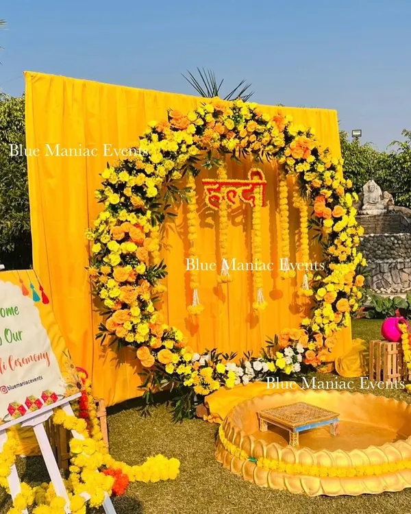 A perfect outdoor Haldi setup. This design features a large floral ring of marigolds against a bright yellow backdrop, with a traditional urli for seating and a custom welcome sign for the ceremony.