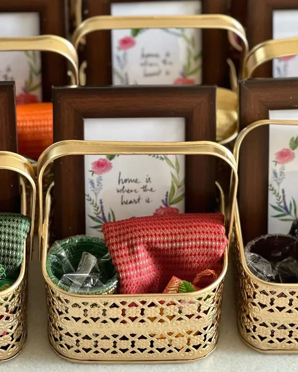 A beautiful housewarming return gift set. Each golden metal basket contains a custom photo frame, a colorful blouse piece, and other traditional items, making it a thoughtful and complete favor.