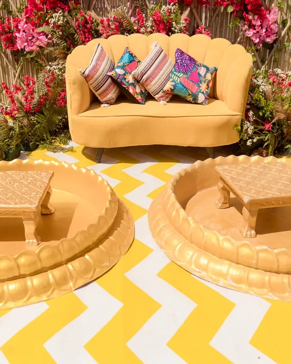 A picture-perfect Haldi seating area. This setup features a bright yellow sofa against a stunning backdrop of a bamboo lattice and a vibrant red bougainvillea arch.