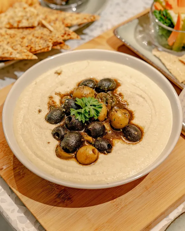 A close-up of our classic hummus, topped with olives and a drizzle of olive oil. Served on a wooden board, it's a simple yet elegant addition to any grazing table.