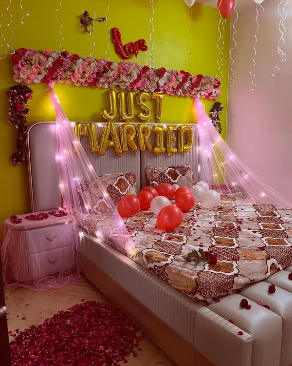 A classic 'Just Married' room decoration to welcome the newlyweds. This setup includes foil balloons, a floral arrangement on the headboard, a canopy with fairy lights, and a floor covered in rose petals.