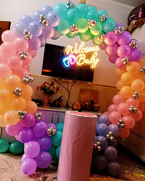 A vibrant and colorful welcome home decoration featuring a circular rainbow balloon arch. The "Welcome Baby" neon sign adds a modern and celebratory touch to the setup.
