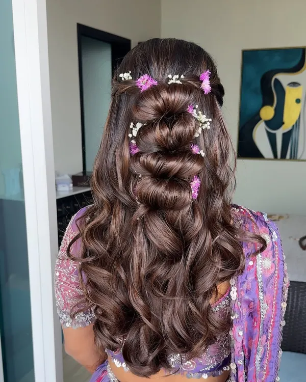 A beautiful half-updo with a twisted bun and soft waves, adorned with lilac and white flowers to match the bride's outfit.