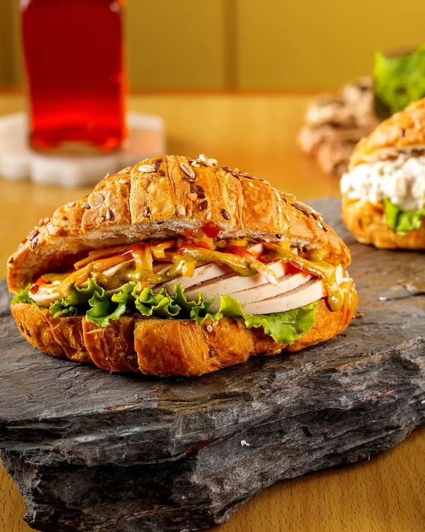 A hearty croissant sandwich filled with fresh ingredients. The rustic stone platter and warm lighting emphasize the sandwich's gourmet quality and the flakiness of the multigrain croissant.