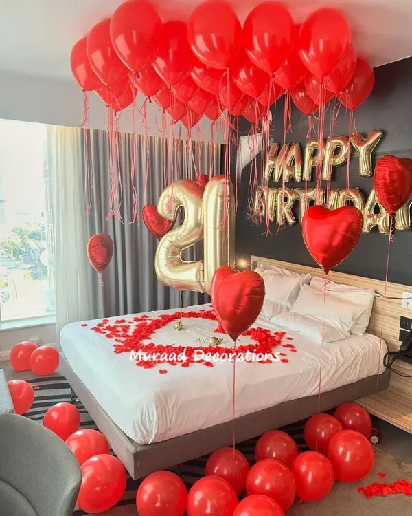 A classic romantic birthday surprise. This room is decorated with red ceiling balloons, 'Happy Birthday' foil letters, large number balloons, and a heart made of rose petals on the bed.