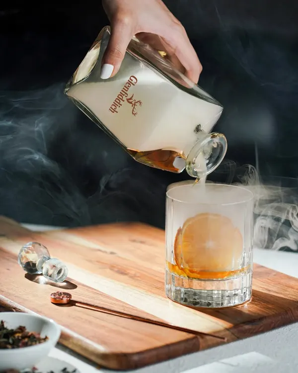 A dramatic pour of smoky whisky into a glass with a perfect ice sphere. This shot uses theatrical smoke and precise lighting to create a luxurious and high-end feel for the brand.