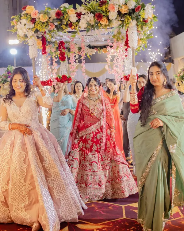A grand bridal entry under a beautiful floral 'phoolon ki chaadar'. The bride looks regal in her heavily embroidered red lehenga, a classic choice for a North Indian wedding ceremony.