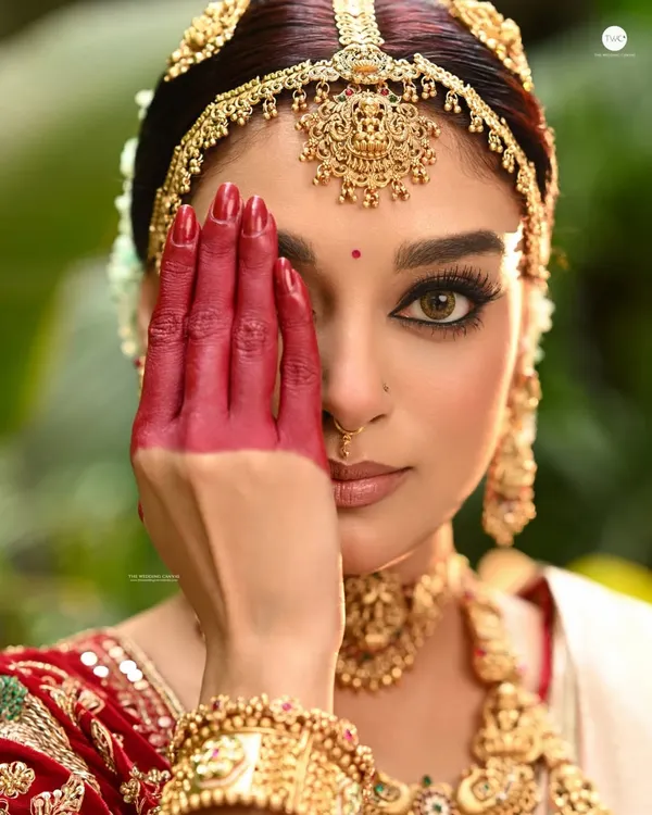 A powerful bridal close-up where the model's hand, decorated with red 'alta', partially covers her face. This creative composition draws attention to her intense eyes and the traditional elements of her look.