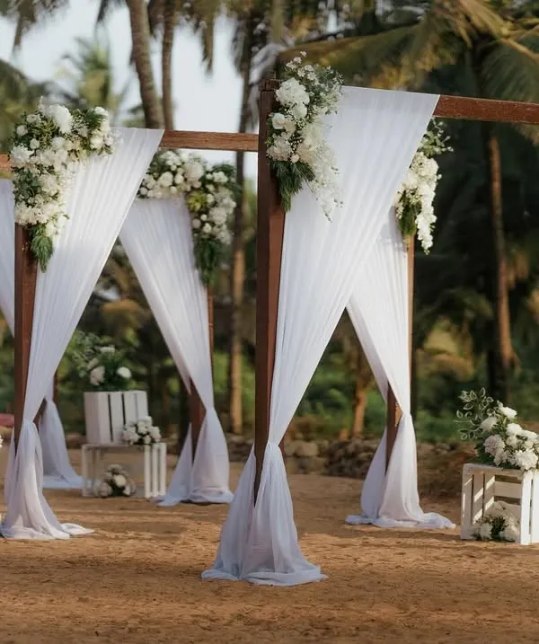 A dreamy beachside wedding setup in Goa. The simple wooden structure with flowing white drapes and florals creates an elegant and romantic aisle right on the sand, with the ocean as the ultimate backdrop.