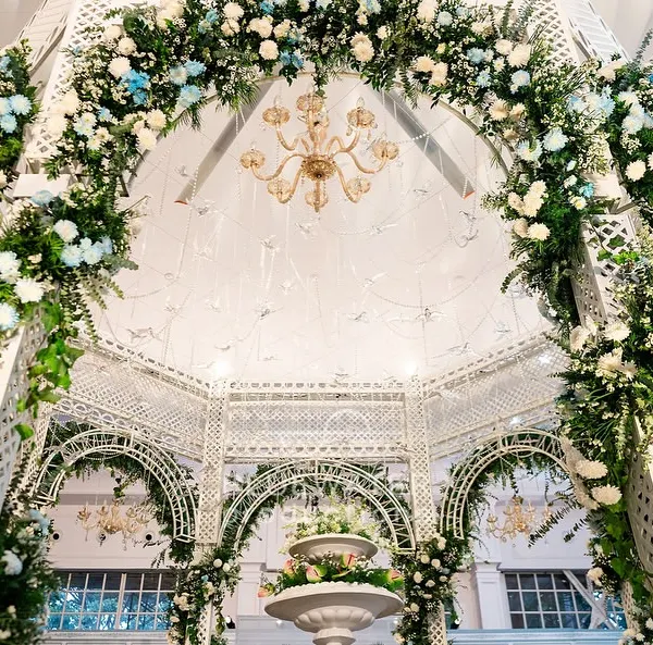 Looking up into a floral arch, where lush greenery and blooms are expertly woven into a white lattice structure, with a chandelier sparkling at the center.