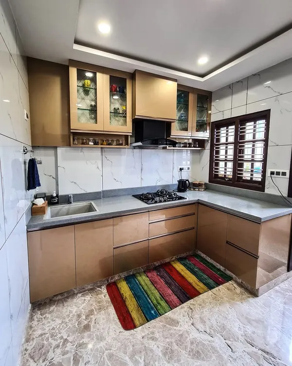 An L-shaped modular kitchen with a sophisticated color palette. The cabinets have a metallic bronze finish, paired with a grey countertop and a white marble-patterned backsplash, creating a modern and luxurious cooking space.