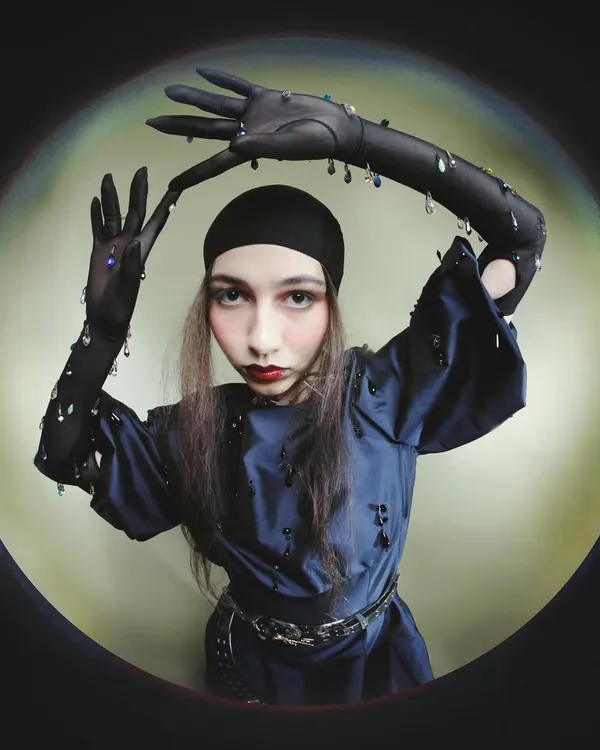 A distorted fisheye portrait from the 'Peek-a-boo' series. The model, wearing embellished gloves and a head covering, frames her face in a playful yet unsettling pose, creating a surreal, peephole effect.