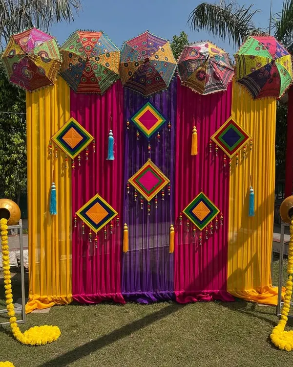 A vibrant photo booth I created for a Mehndi function. It features a backdrop of colorful drapes, Rajasthani umbrellas, and traditional hangings, with decorated pots adding a rustic touch.