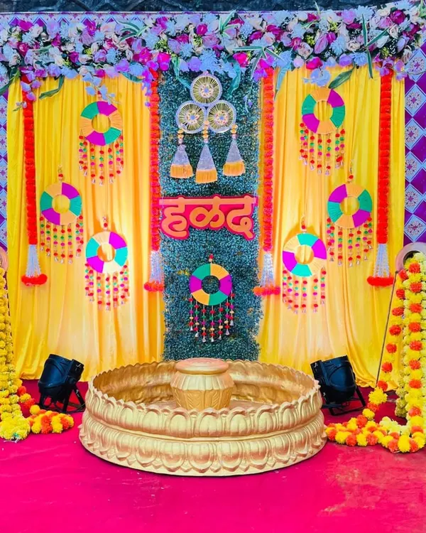 A vibrant Haldi ceremony backdrop featuring bright yellow curtains and a central panel of artificial grass. The setup is decorated with colorful hanging ornaments, marigold garlands, and a 'हळद' sign, creating a festive atmosphere.