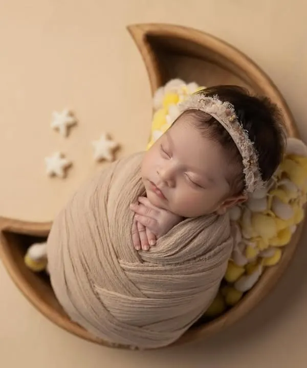 A close up of a newborn swaddled in a neutral wrap, resting in a wooden moon bowl. The yellow floral details and tiny stars add a touch of whimsy to this dreamy setup.