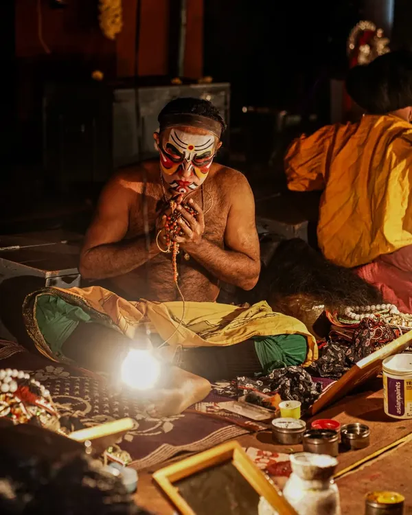 An intimate look at a Yakshagana artist in the 'Chowki' as he meticulously applies his makeup. This close-up shot captures the intense focus and ritualistic nature of the transformation process before the performance begins.