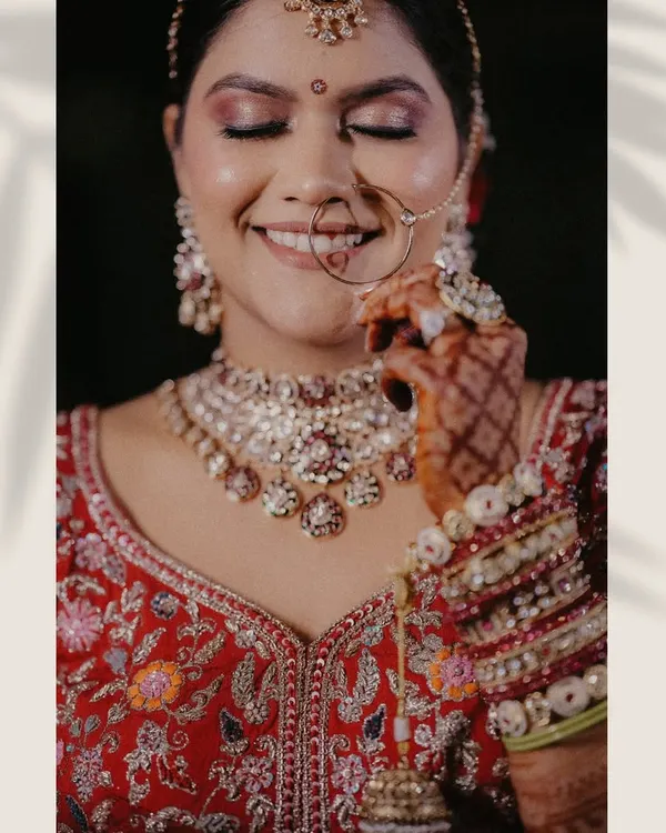 A stunning close-up of the bride, highlighting her intricate jewelry, flawless makeup, and the rich embroidery of her red lehenga. This is a perfect example of a fine art bridal portrait.